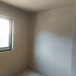 Wnętrze pokoju po nałożeniu gładzi, widoczne świeżo otynkowane ściany w odcieniach beżu i szarości, okno z widocznym światłem dziennym oraz surowy betonowy podkład podłogowy.