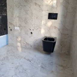 Zamontowana, czarna miska bidetowa wisząca na ścianie wyłożonej jasnymi płytkami imitującymi marmur, z widocznymi otworami na armaturę i czarnym panelem sterującym spłuczką.