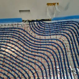 Niebieskie rury ogrzewania podłogowego układane na srebrnej folii z siatką, mocowane czarnymi klipsami. Widoczne białe rury i otwory w suficie z wystającymi kablami.