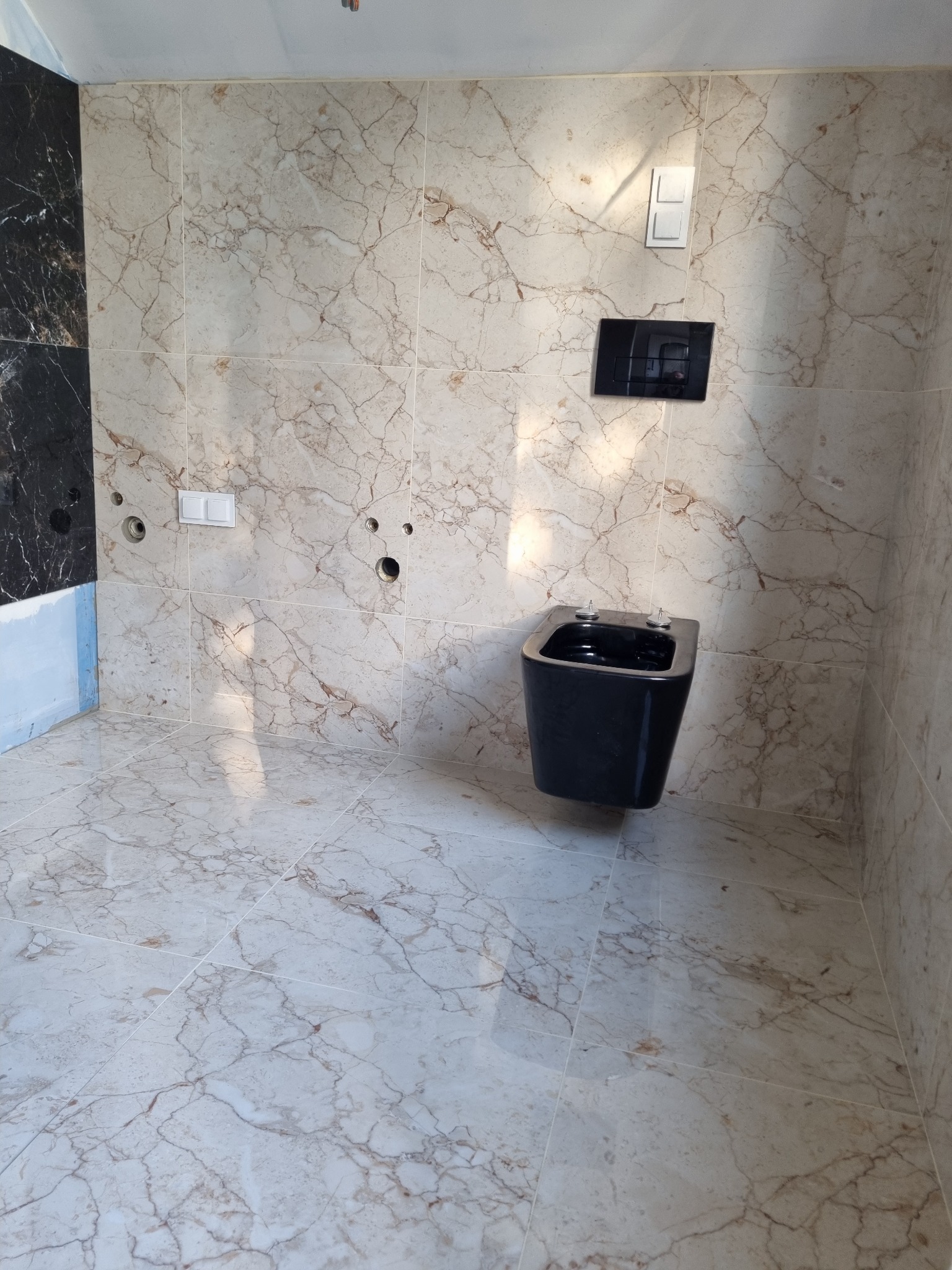 Zamontowana, czarna miska bidetowa wisząca na ścianie wyłożonej jasnymi płytkami imitującymi marmur, z widocznymi otworami na armaturę i czarnym panelem sterującym spłuczką.