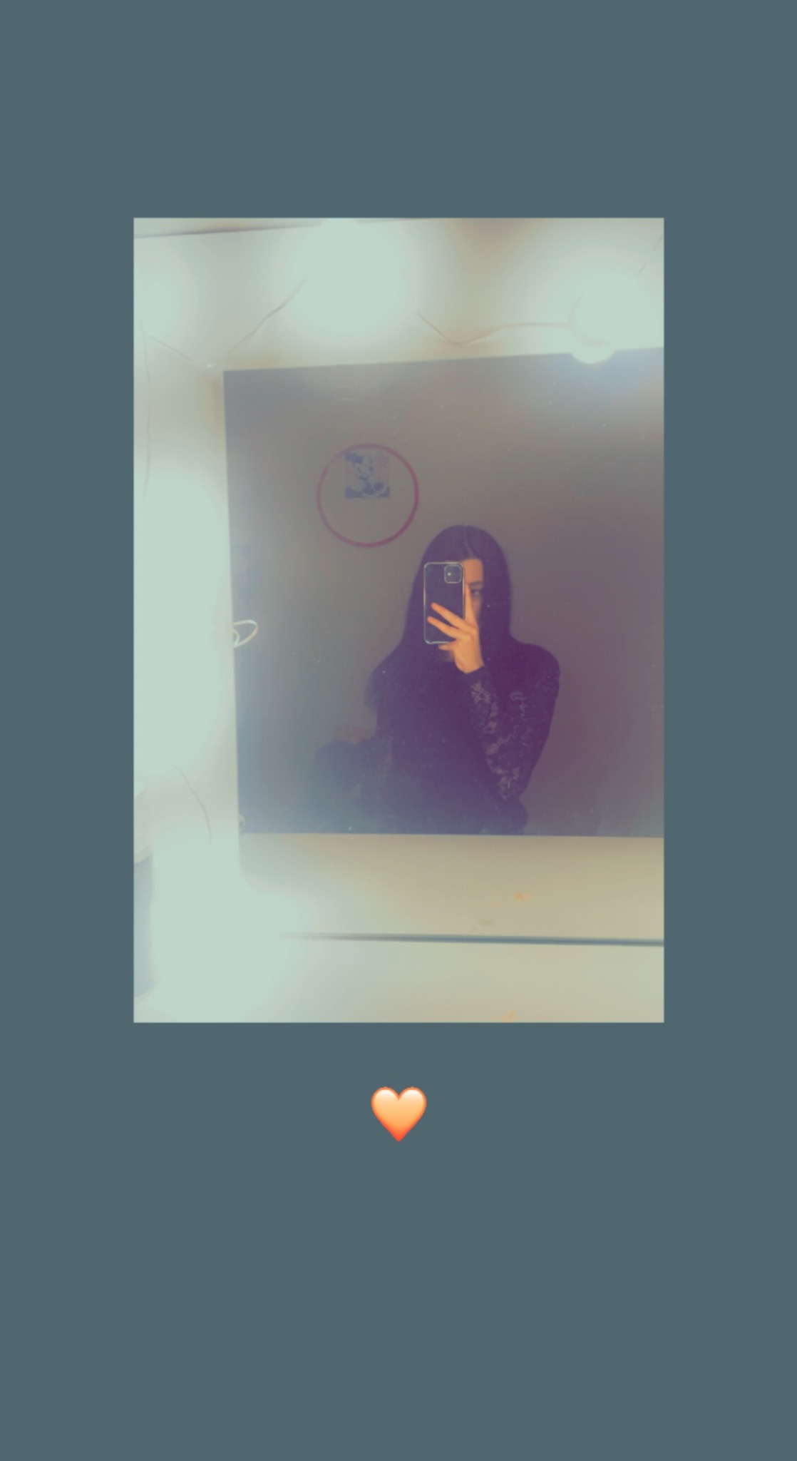 Odbicie kobiety robiącej selfie w lustrze łazienkowym z oświetleniem, widoczna naklejka na ścianie i umywalka.