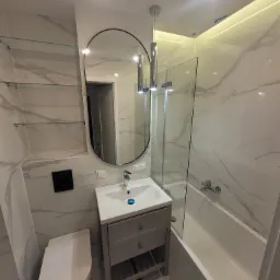 Nowoczesna łazienka z białymi marmurkowymi płytkami, owalnym lustrem z czarną ramą, wiszącą szafką pod umywalką w kolorze szarym, szklanymi półkami i kabiną prysznicową z pojedynczymi drzwiami.