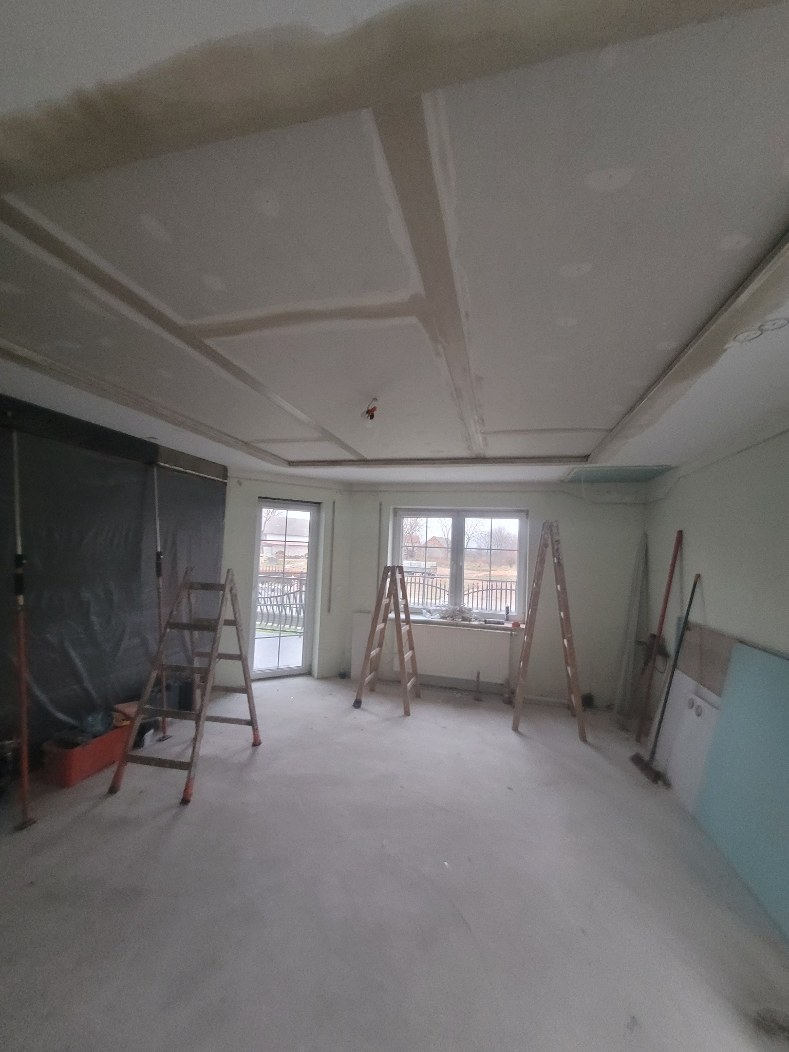 Wnętrze w trakcie prac wykończeniowych, widoczne płyty kartonowo-gipsowe na suficie z dekoracyjnymi wgłębieniami, drabiny, okno z widokiem na podwórze i częściowo zasłonięte okno po lewej stronie.