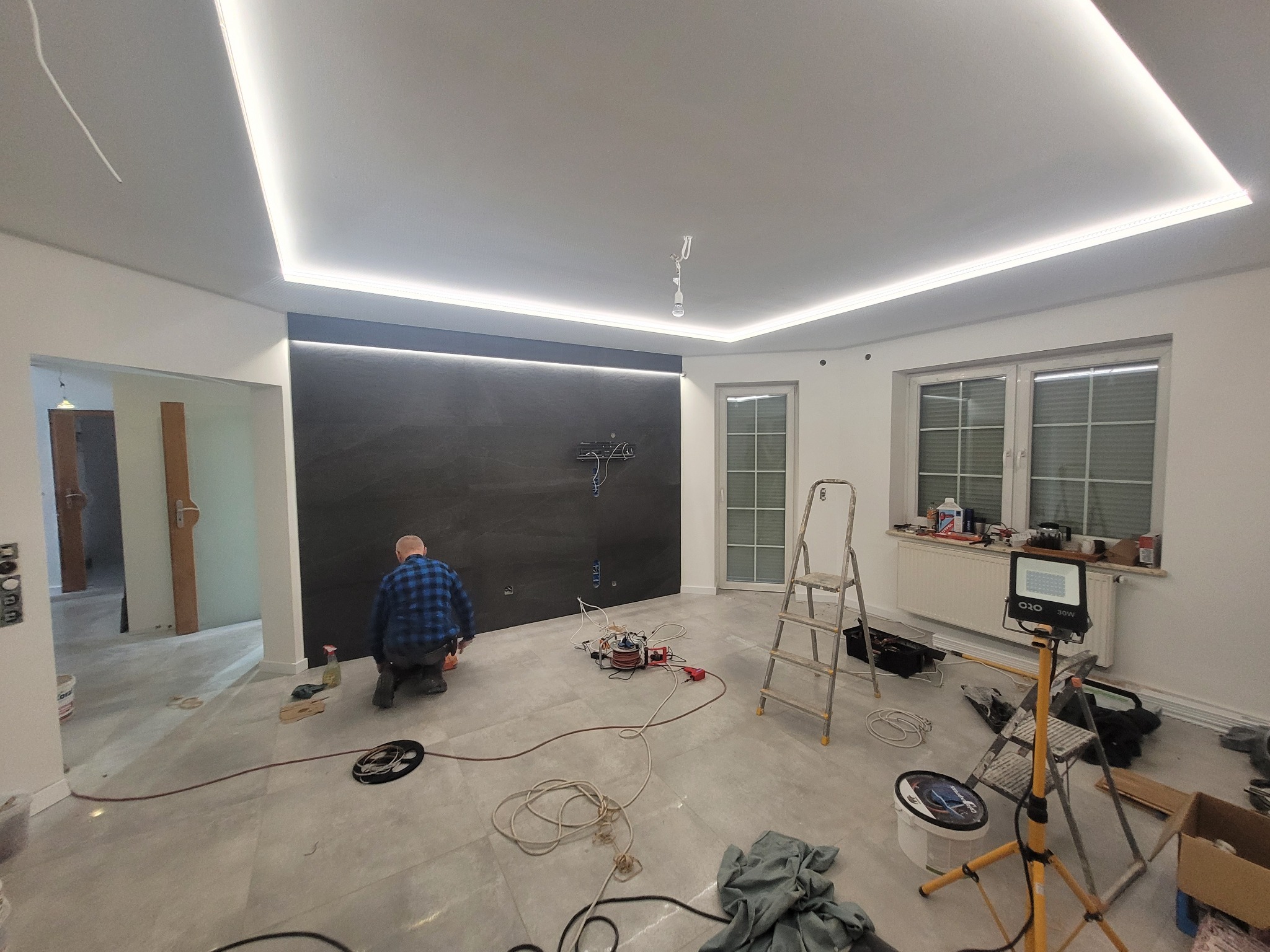 Pokój w trakcie remontu z widocznym pracownikiem w niebieskiej koszuli, stojąca drabina, lampa oświetleniowa na statywie i przewody elektryczne na podłodze. Widoczny fragment ściany pomalowanej...