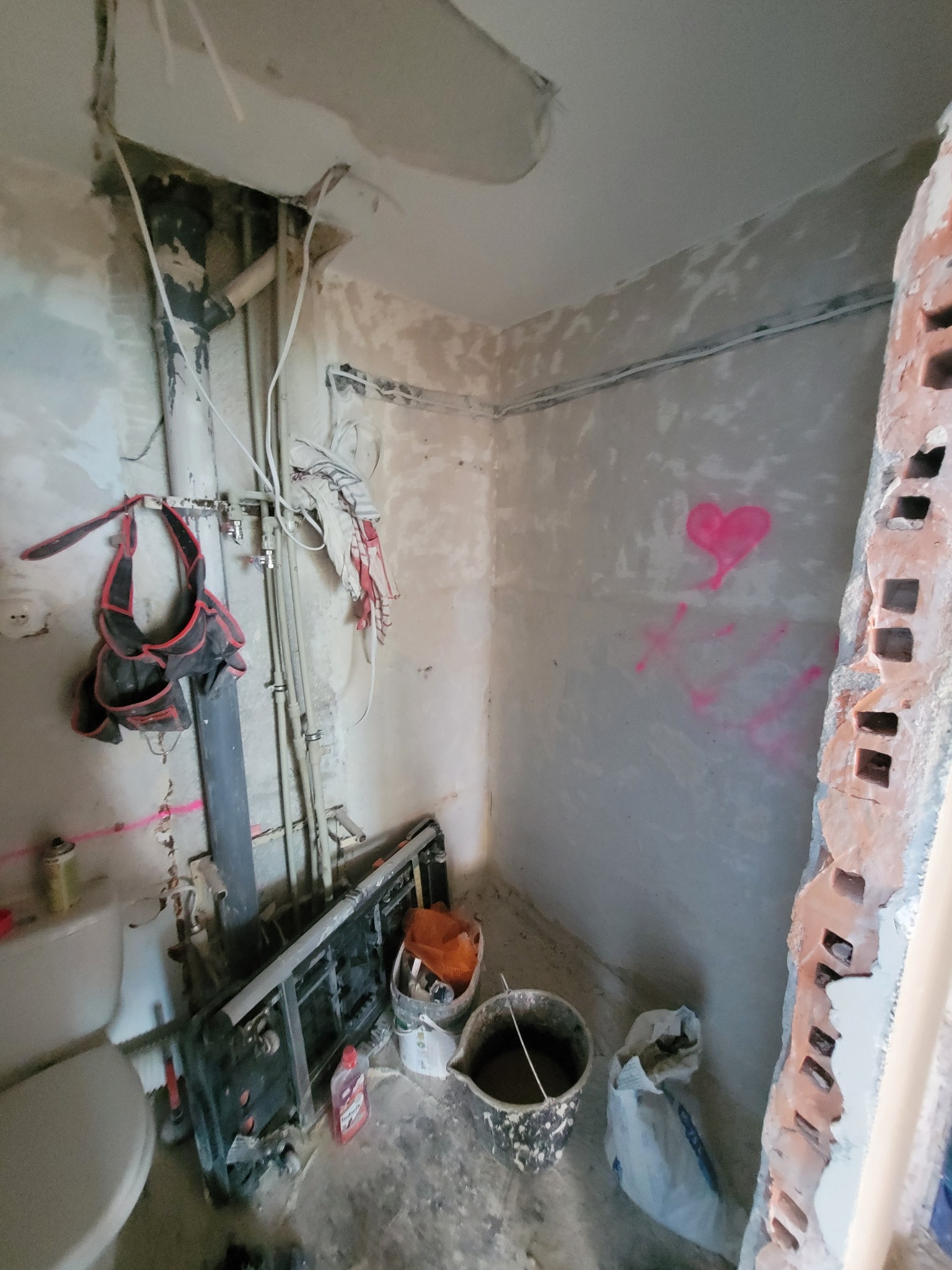 Wnętrze pomieszczenia w trakcie prac remontowych: widoczne odsłonięte rury, wiszący pas narzędziowy, fragment ściany z różowym sercem namalowanym sprayem, pozostałości po skuciu płytek i fragment...