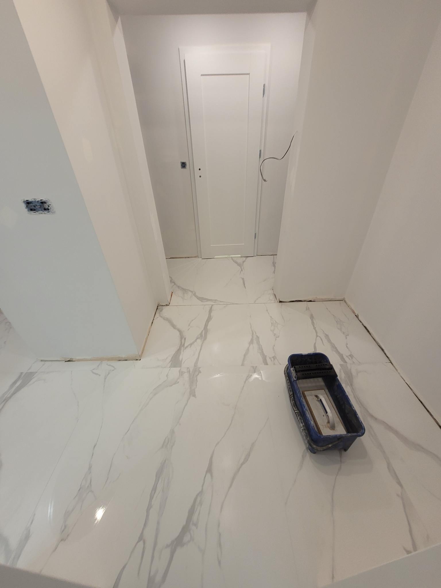 Biała marmurowa podłoga w korytarzu po ułożeniu płytek, widoczny pojemnik z narzędziami do fugowania, surowy stan ścian i brak listew przypodłogowych.