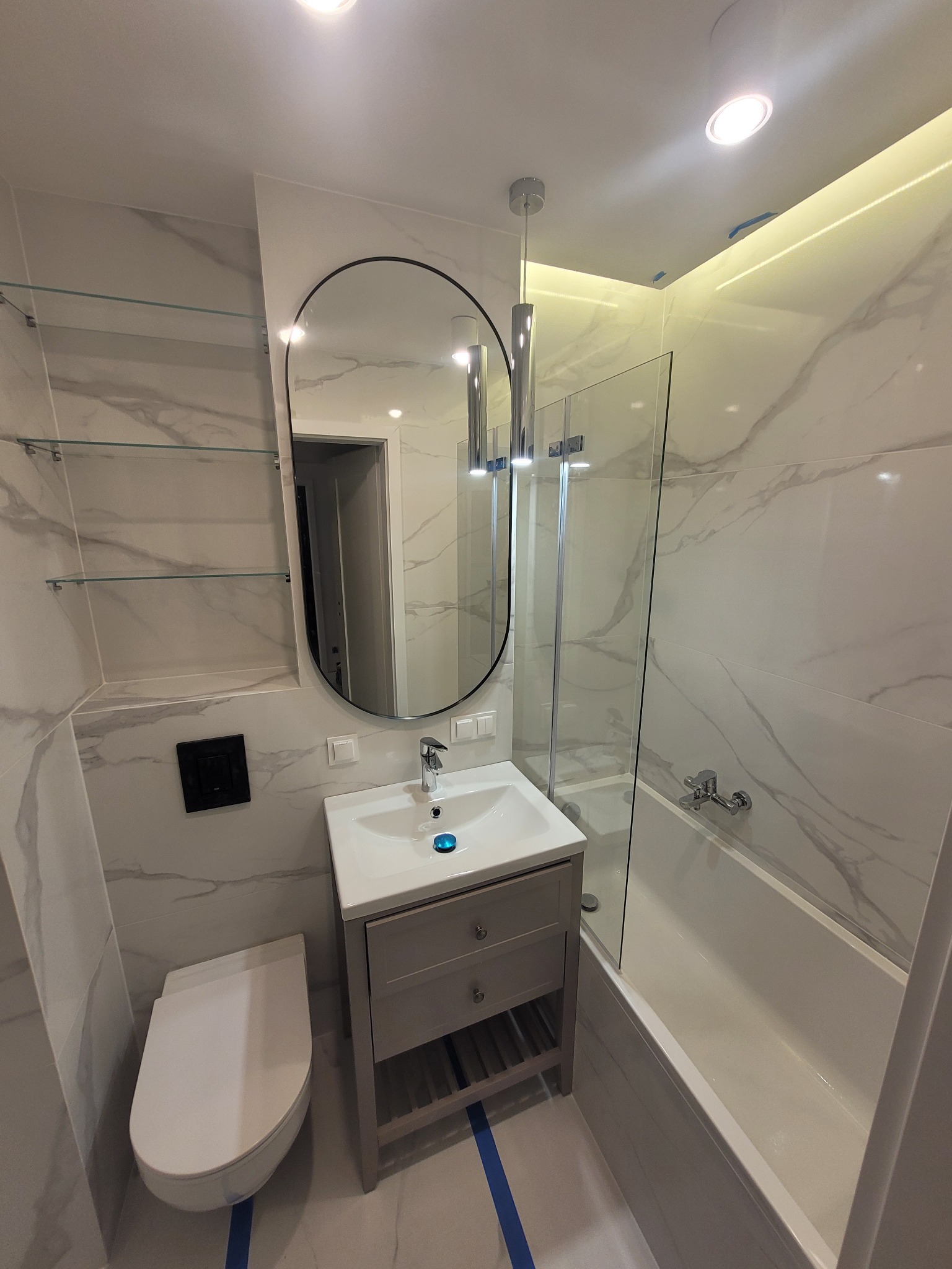 Nowoczesna łazienka z białymi marmurkowymi płytkami, owalnym lustrem z czarną ramą, wiszącą szafką pod umywalką w kolorze szarym, szklanymi półkami i kabiną prysznicową z pojedynczymi drzwiami.