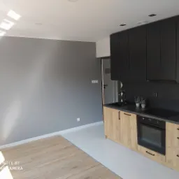 Nowoczesna kuchnia z czarnymi górnymi szafkami, drewnopodobnymi dolnymi szafkami, heksagonalnymi płytkami na ścianie i szarą ścianą w tle. Część podłogi wyłożona panelami, a część płytkami.