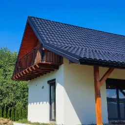 Nowoczesny dom z balkonem z czarnymi metalowymi balustradami i drewnianymi elementami konstrukcyjnymi, na tle zieleni i błękitnego nieba.