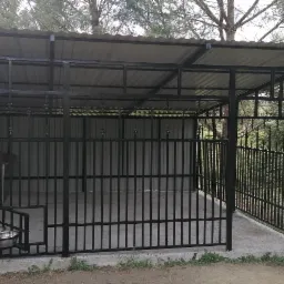 Metalowa konstrukcja kojca dla psa z zadaszeniem, miskami na karmę i budą wewnątrz, otoczona zielenią.