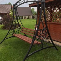 RAF-SPAW RAFAŁ KOPCZYK - Metalowa huśtawka ogrodowa z drewnianym siedziskiem, ozdobiona motywami roślinnymi, ustawiona na trawniku przed drewnianą altaną.