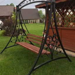 Metalowa huśtawka ogrodowa z drewnianym siedziskiem, ozdobiona motywami roślinnymi, ustawiona na trawniku przed drewnianą altaną.