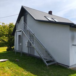 RAF-SPAW RAFAŁ KOPCZYK - Zewnętrzne, metalowe schody z balustradą prowadzące do drzwi na piętrze domu. Widoczne elementy konstrukcyjne i mocowania. Dom z szarą blachodachówką.