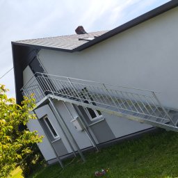 RAF-SPAW RAFAŁ KOPCZYK - Zewnętrzne schody metalowe z balustradą prowadzące do drzwi na piętrze budynku. Widok z dołu, zieleń w tle. Konstrukcja ocynkowana, nowoczesny design.