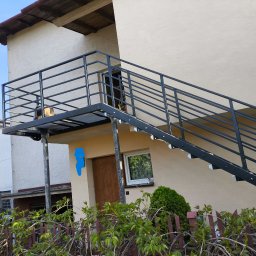 RAF-SPAW RAFAŁ KOPCZYK - Zewnętrzne schody metalowe z balustradą, prowadzące do drzwi na piętrze budynku. Konstrukcja pomalowana na czarno, widok z dołu, zieleń w tle.