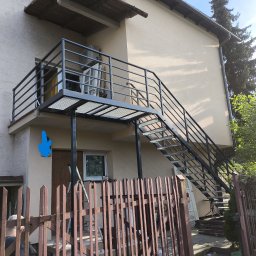 RAF-SPAW RAFAŁ KOPCZYK - Zewnętrzne schody metalowe z balustradą prowadzące na balkon domu jednorodzinnego. Konstrukcja w kolorze czarnym, ażurowe stopnie. Widok z dołu, częściowo zasłonięte przez płot.