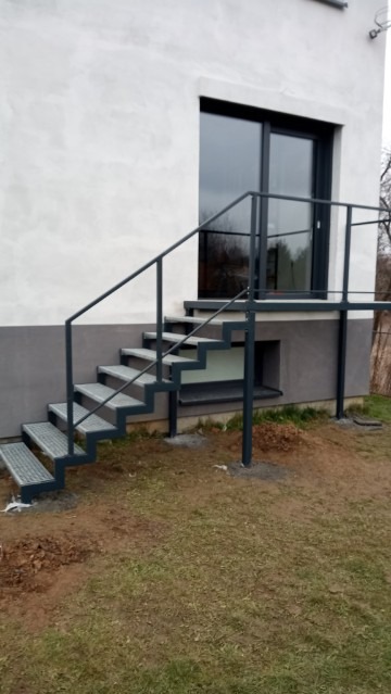 Zewnętrzne, ocynkowane schody z metalu prowadzące do drzwi balkonowych na podwyższeniu, z szarą balustradą, na tle białego budynku.