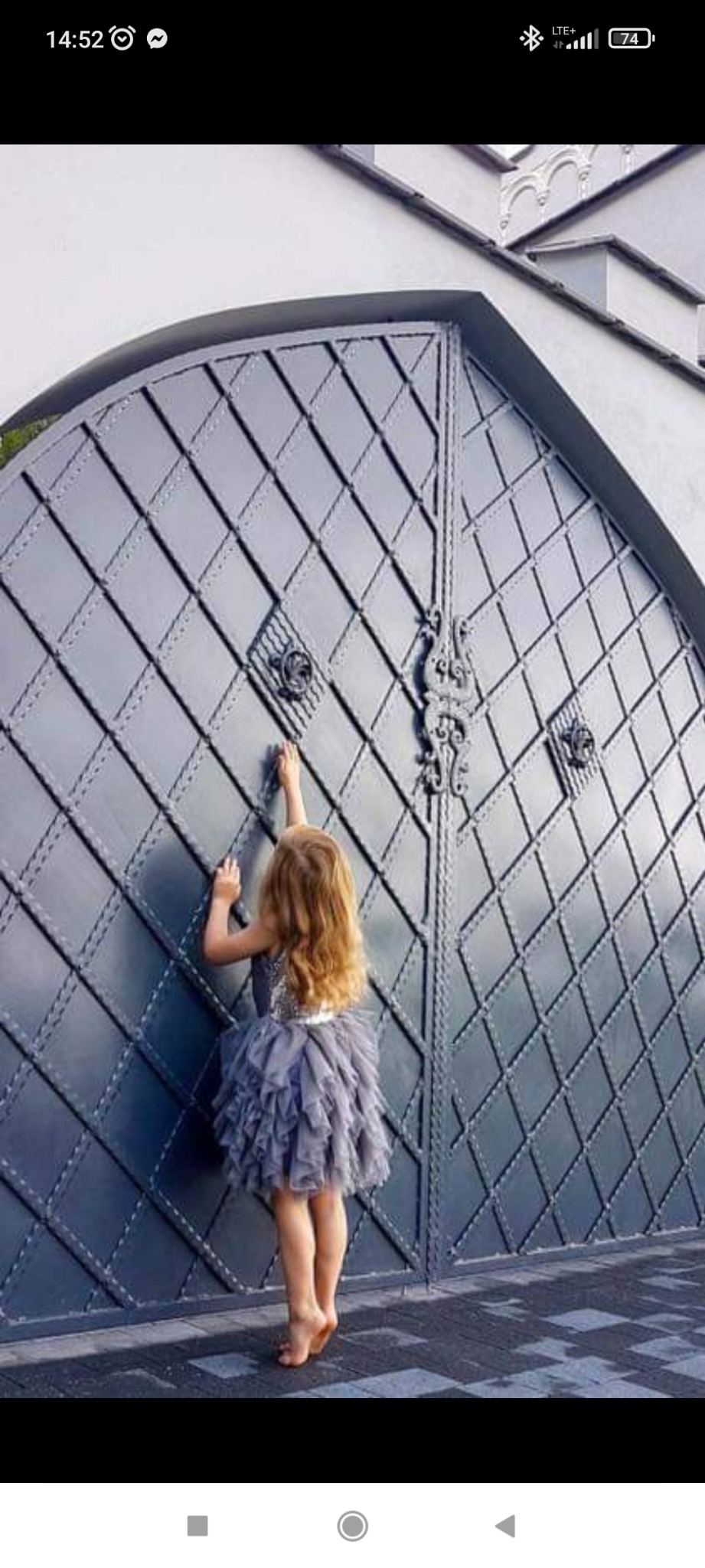 Dziewczynka w tiulowej sukience sięga do ozdobnej metalowej bramy z romboidalnym wzorem i kutymi detalami.