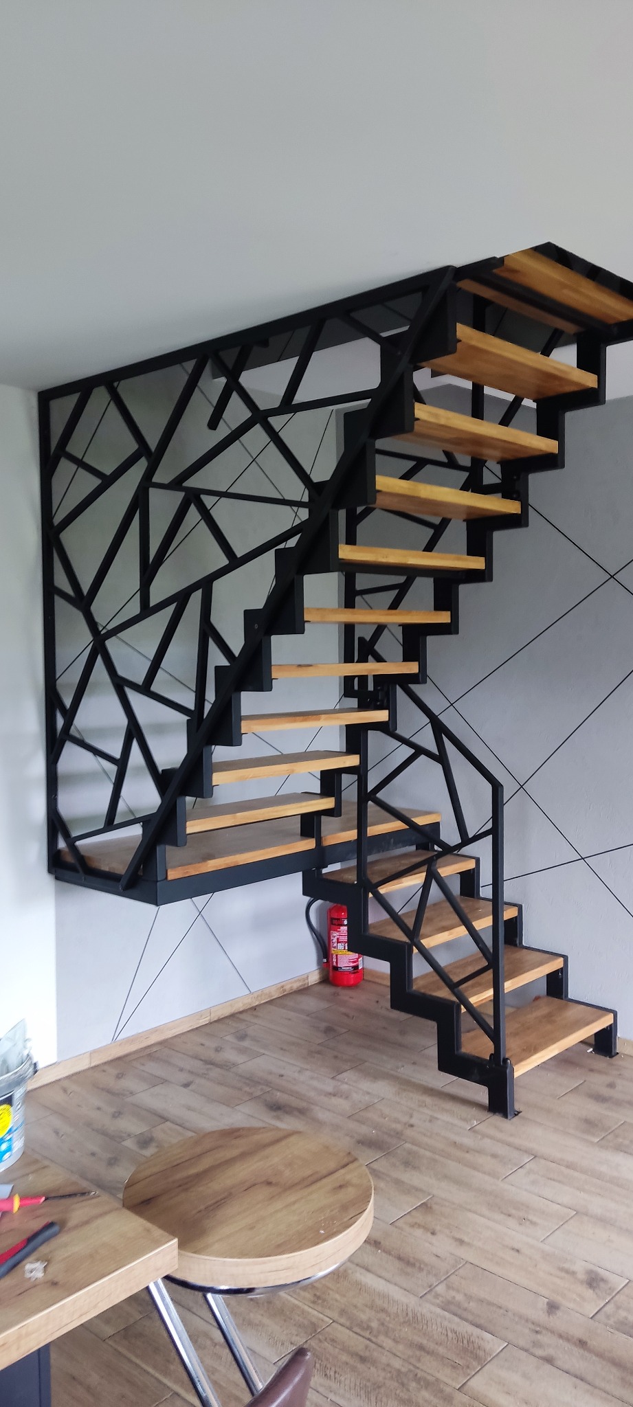 Nowoczesne schody z metalową konstrukcją w geometryczne wzory i drewnianymi stopniami, widok na fragment pomieszczenia z gaśnicą i stołkiem barowym.