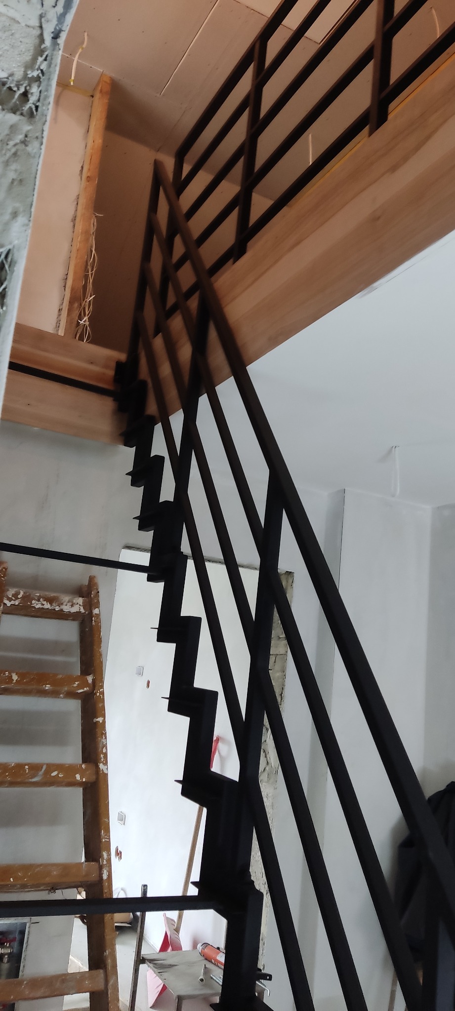 Czarne metalowe schody z minimalistyczną balustradą w surowym wnętrzu, widoczna drewniana konstrukcja stropu i drabina malarska.