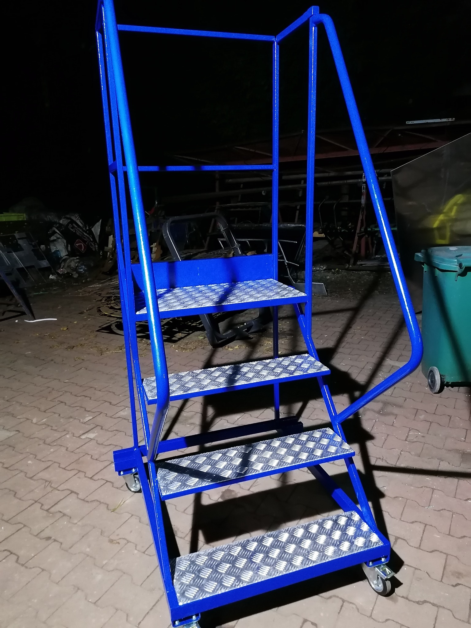 Niebieskie, metalowe schody robocze na kółkach z antypoślizgowymi stopniami, sfotografowane na zewnątrz w słabym oświetleniu.