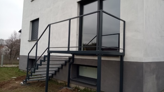 Zewnętrzne schody metalowe z balustradą, prowadzące do drzwi balkonowych w nowoczesnym domu, widoczne z boku, z elementami ocynkowanej stali.