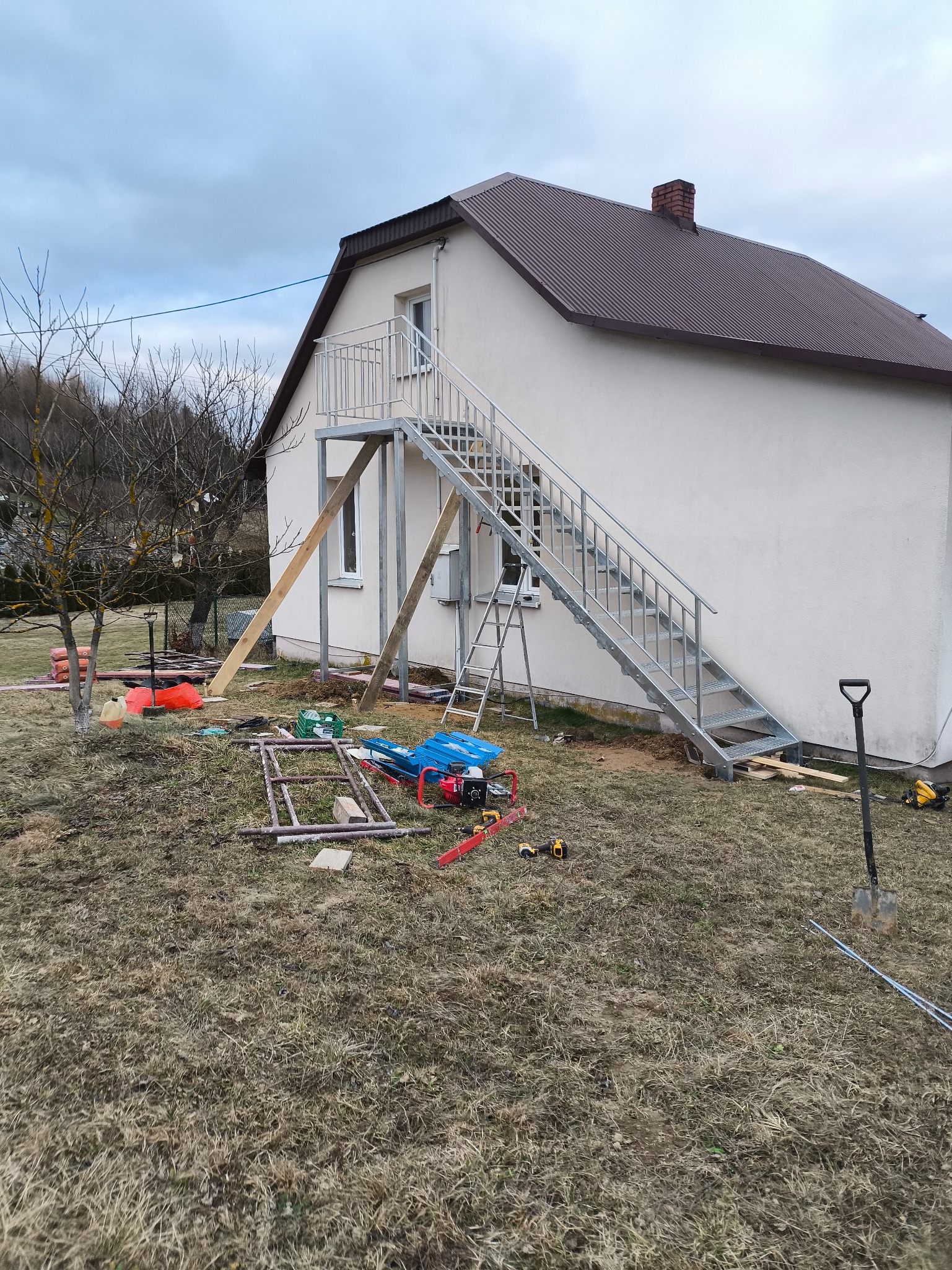 Zewnętrzne, metalowe schody z balustradą prowadzące do drzwi na piętrze domu, widoczne narzędzia i materiały budowlane na trawniku.