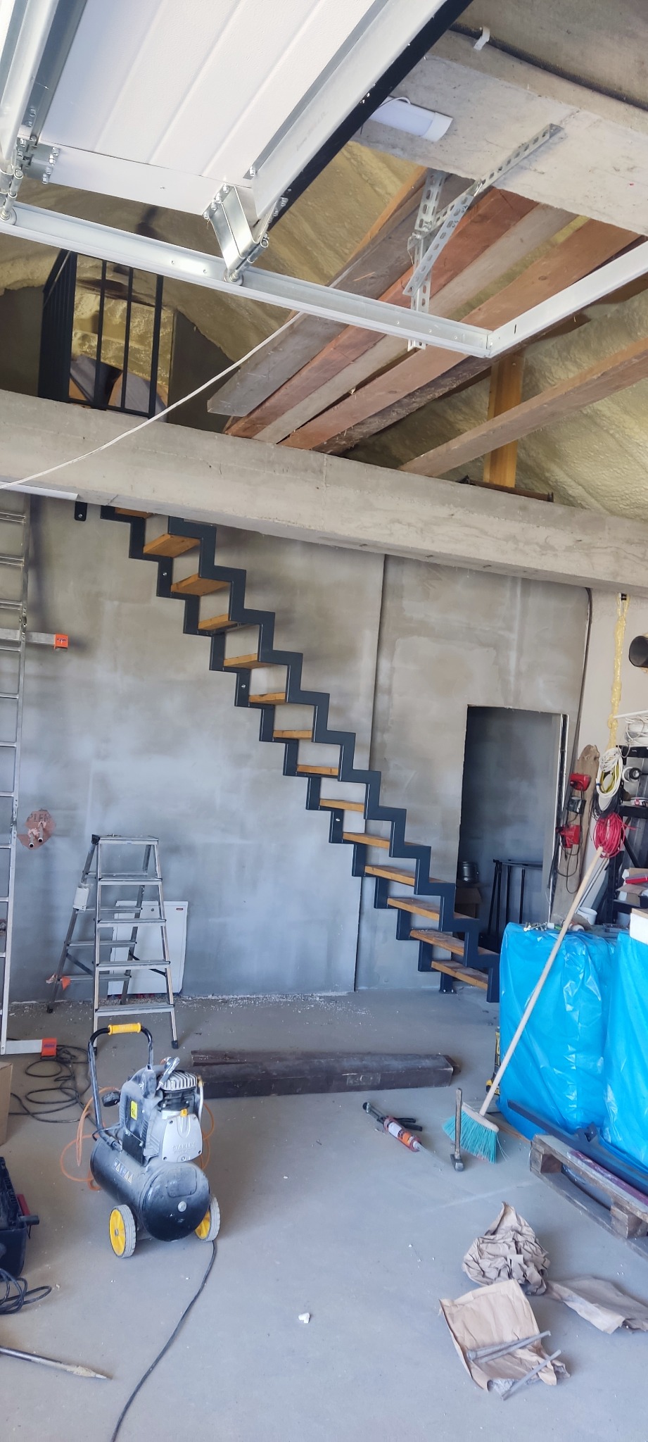 Wewnętrzne schody wspornikowe z metalową konstrukcją i drewnianymi stopniami w surowym wnętrzu garażu, widoczny kompresor i drabina.