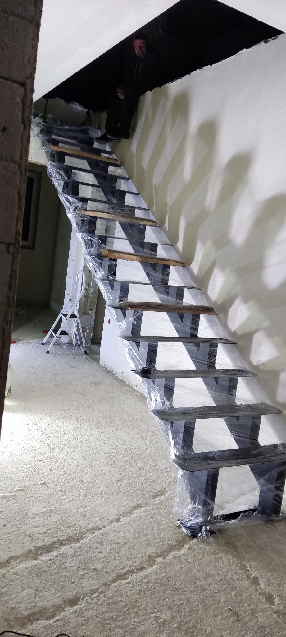 Nowe schody z metalową konstrukcją i drewnianymi stopniami, zabezpieczone folią ochronną podczas prac wykończeniowych; w tle widoczna drabina i osoba.