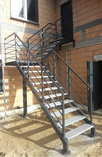 Zewnętrzne schody metalowe z perforowanymi stopniami prowadzące do drzwi na piętrze w surowym stanie budynku, widać świeże ślady spawania.