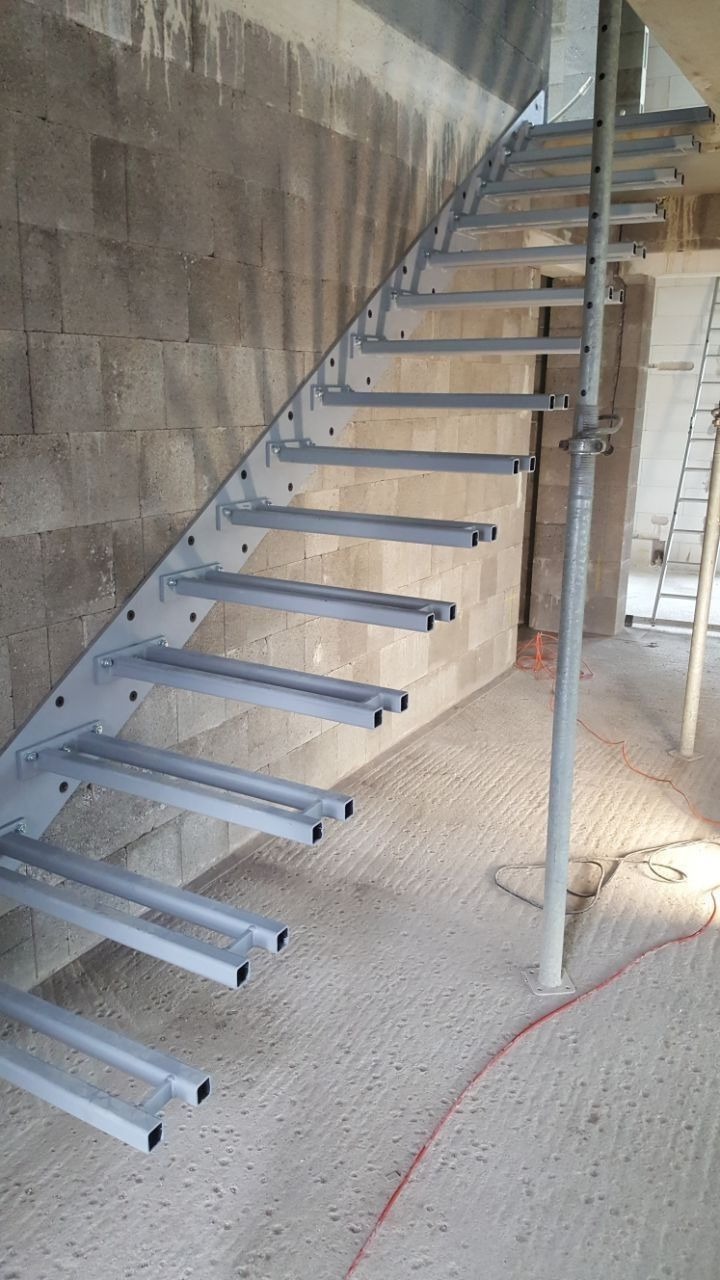 Szare, metalowe schody spiralne bez stopni, przymocowane do surowej ściany z betonowych bloczków, widoczne elementy konstrukcyjne i podpora tymczasowa.