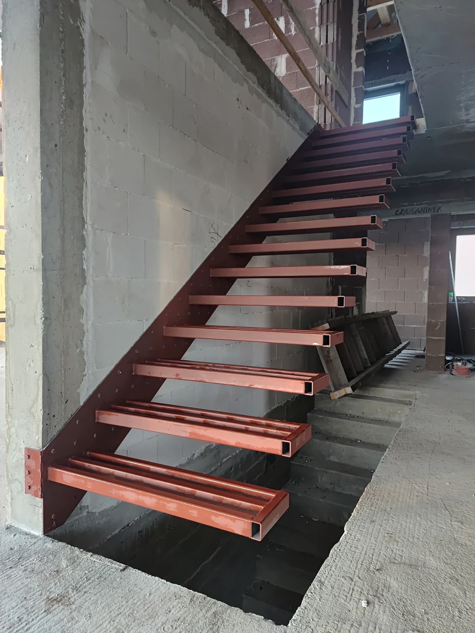 Surowe, metalowe schody o konstrukcji wspornikowej, z rur prostokątnych pomalowanych na rdzawy kolor, zamontowane w surowym stanie budynku, widoczne bloczki betonowe i drabina.