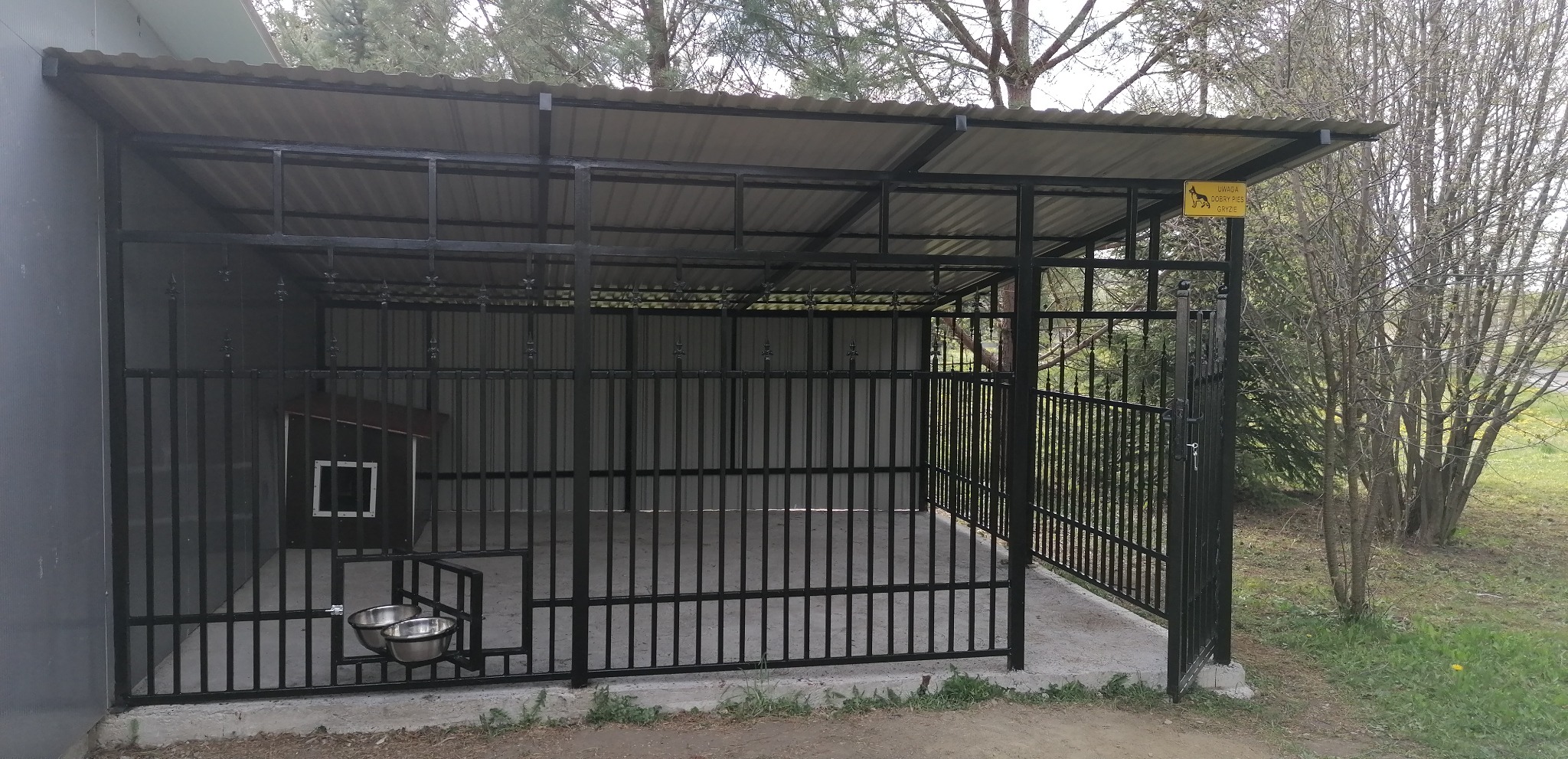 Metalowa konstrukcja kojca dla psa z zadaszeniem, miskami na karmę i budą wewnątrz, otoczona zielenią.