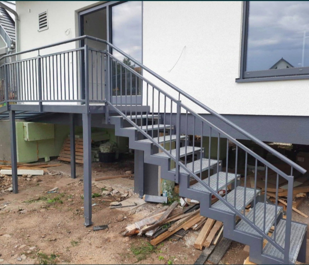 Zewnętrzne schody z metalu pomalowane na szaro, prowadzące do drzwi balkonowych, z widocznymi elementami konstrukcyjnymi i drewnianymi odpadami budowlanymi na ziemi.