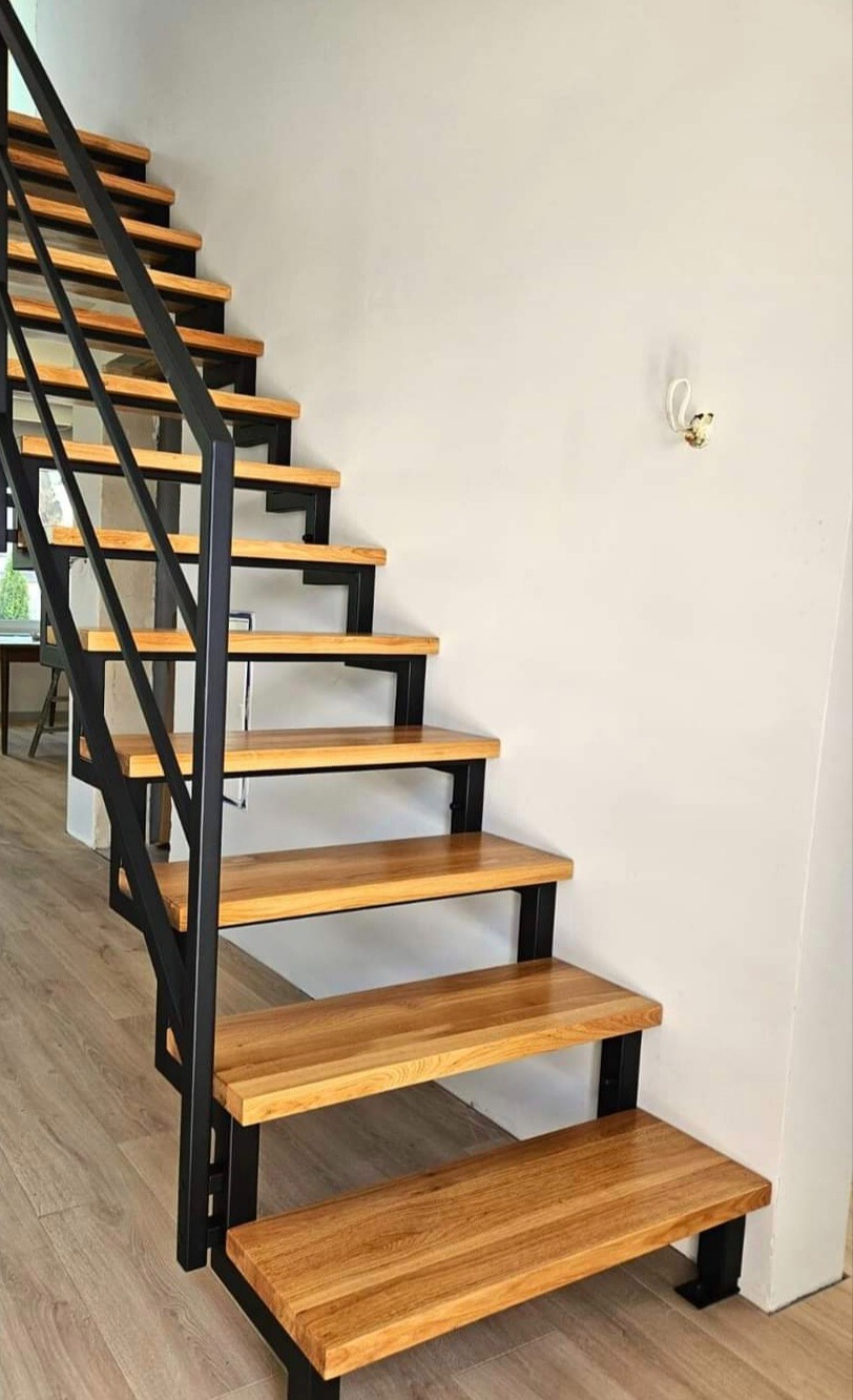 Nowoczesne schody o drewnianych stopniach i czarnej, metalowej konstrukcji, w jasnym wnętrzu.