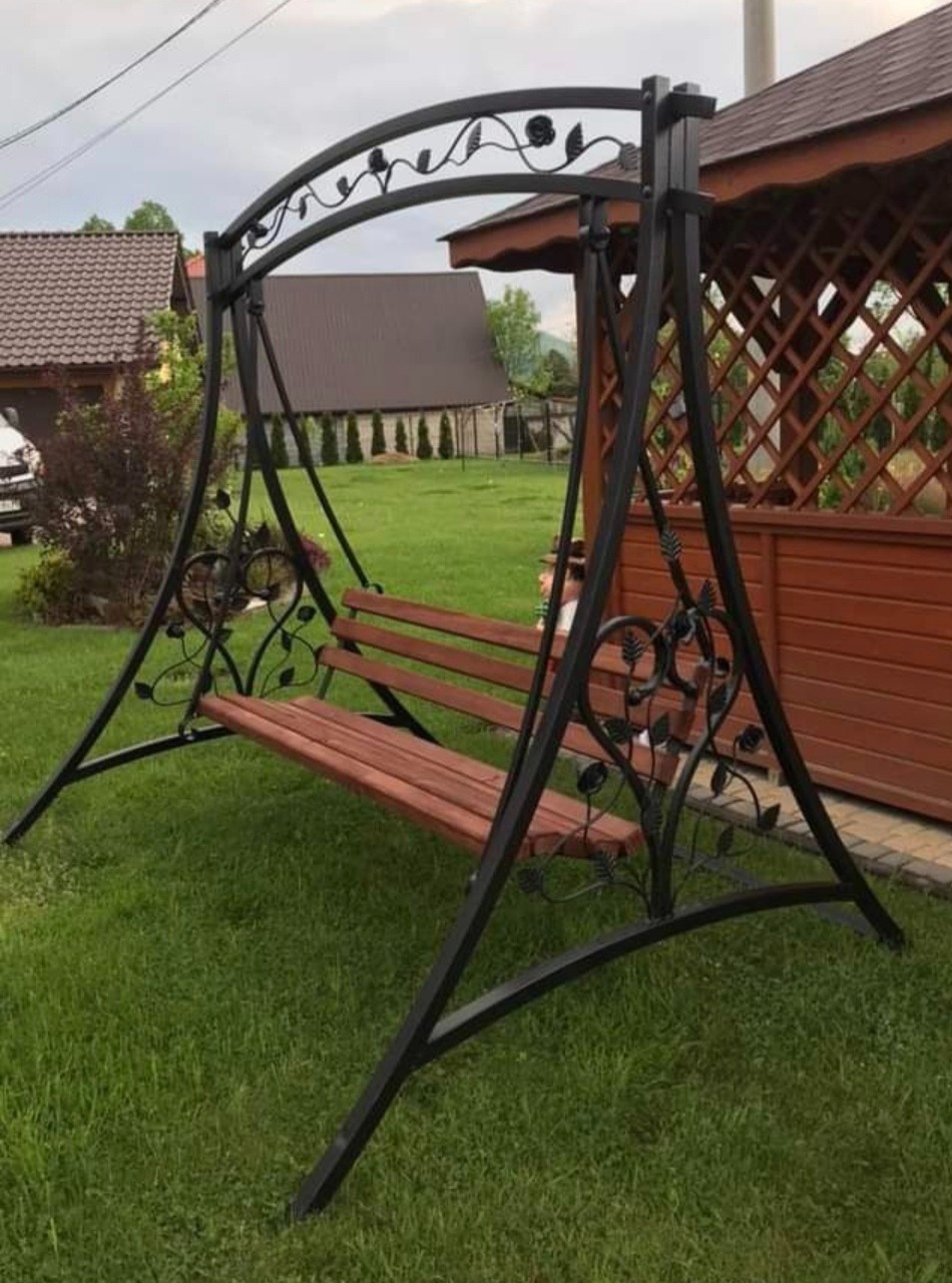 Metalowa huśtawka ogrodowa z drewnianym siedziskiem, ozdobiona motywami roślinnymi, ustawiona na trawniku przed drewnianą altaną.