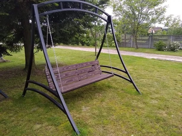 Metalowa huśtawka ogrodowa z drewnianym siedziskiem, zawieszona na łańcuchach, ustawiona na trawniku przed domem.