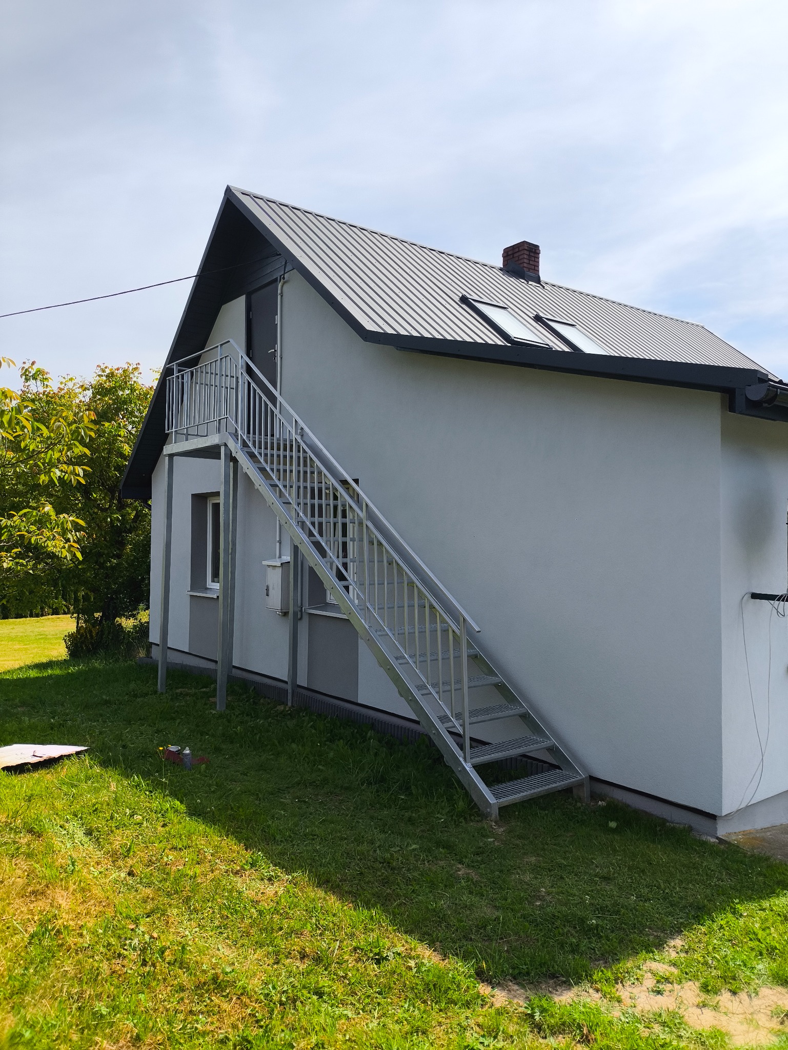 Zewnętrzne, metalowe schody z balustradą prowadzące do drzwi na piętrze domu. Widoczne elementy konstrukcyjne i mocowania. Dom z szarą blachodachówką.