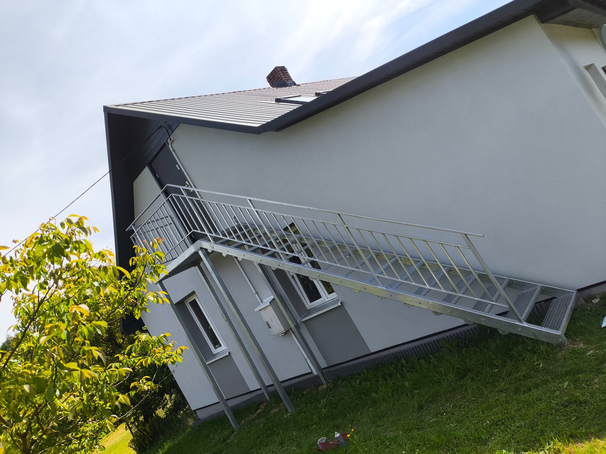 Zewnętrzne schody metalowe z balustradą prowadzące do drzwi na piętrze budynku. Widok z dołu, zieleń w tle. Konstrukcja ocynkowana, nowoczesny design.
