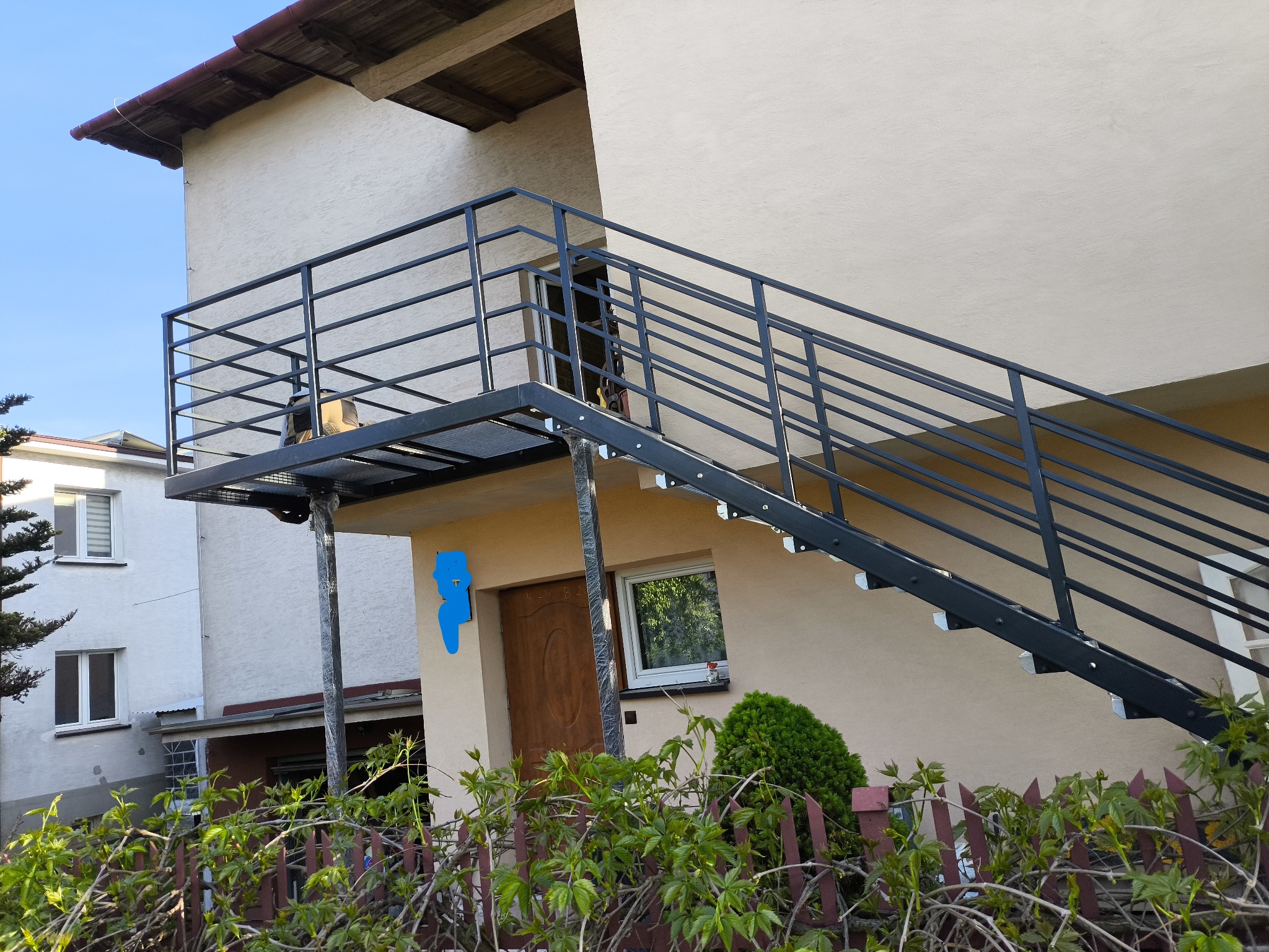 Zewnętrzne schody metalowe z balustradą, prowadzące do drzwi na piętrze budynku. Konstrukcja pomalowana na czarno, widok z dołu, zieleń w tle.