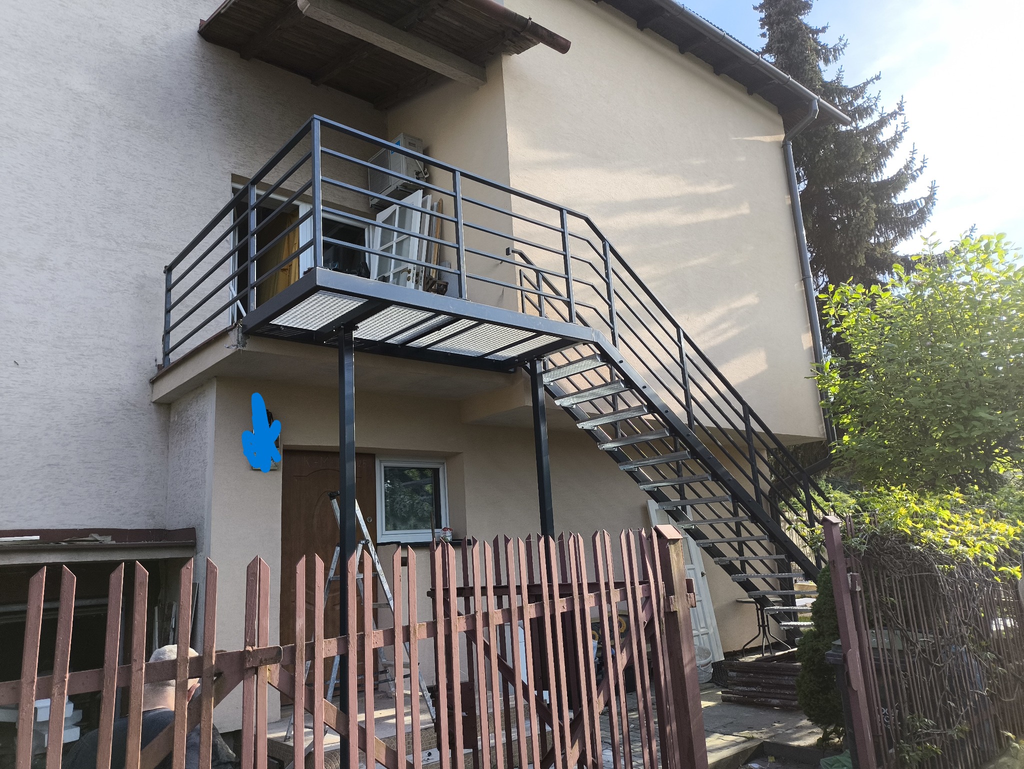 Zewnętrzne schody metalowe z balustradą prowadzące na balkon domu jednorodzinnego. Konstrukcja w kolorze czarnym, ażurowe stopnie. Widok z dołu, częściowo zasłonięte przez płot.
