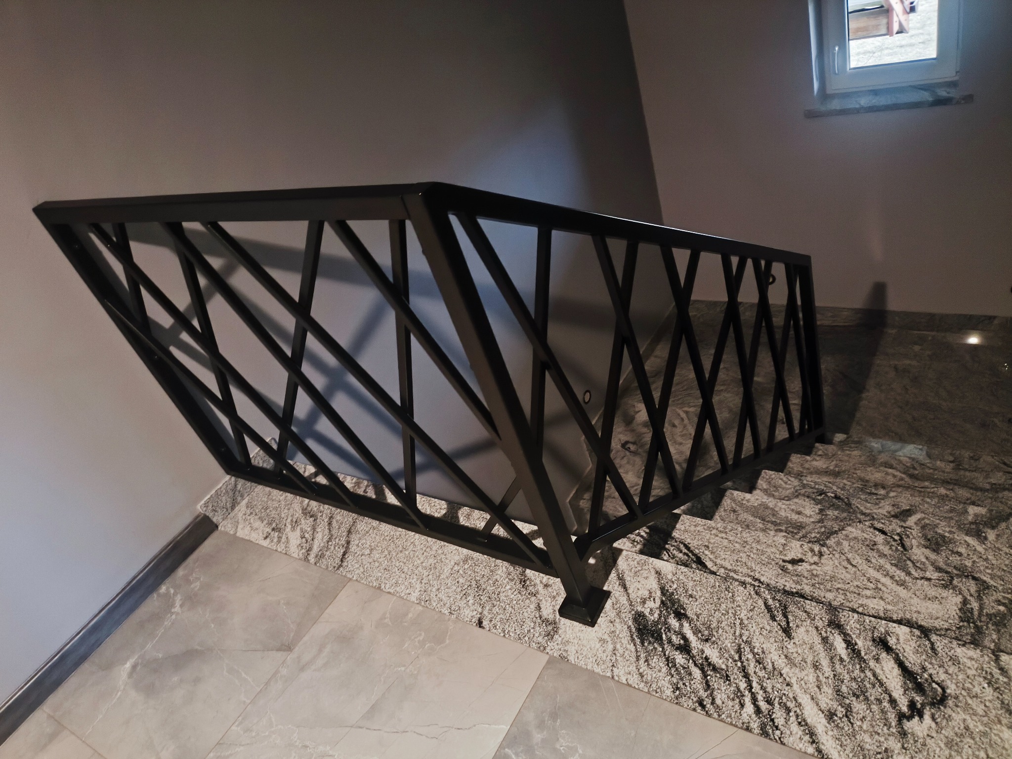 Nowoczesna, czarna balustrada schodowa z geometrycznym wzorem, zamontowana na marmurowych schodach. Widok z góry, oświetlenie naturalne z okna w tle.