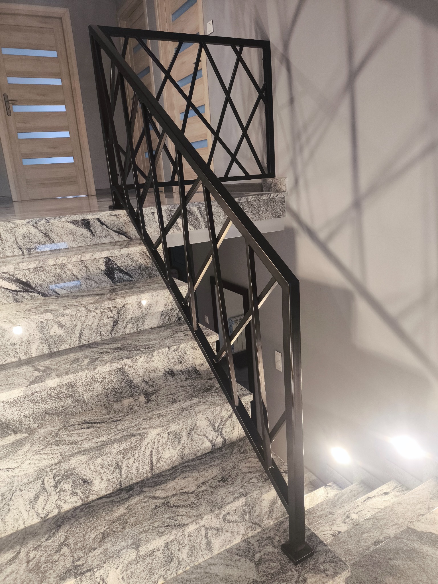 Nowoczesna, czarna balustrada metalowa o geometrycznym wzorze na schodach z szarego, wzorzystego kamienia. Widok z dołu, oświetlenie punktowe.
