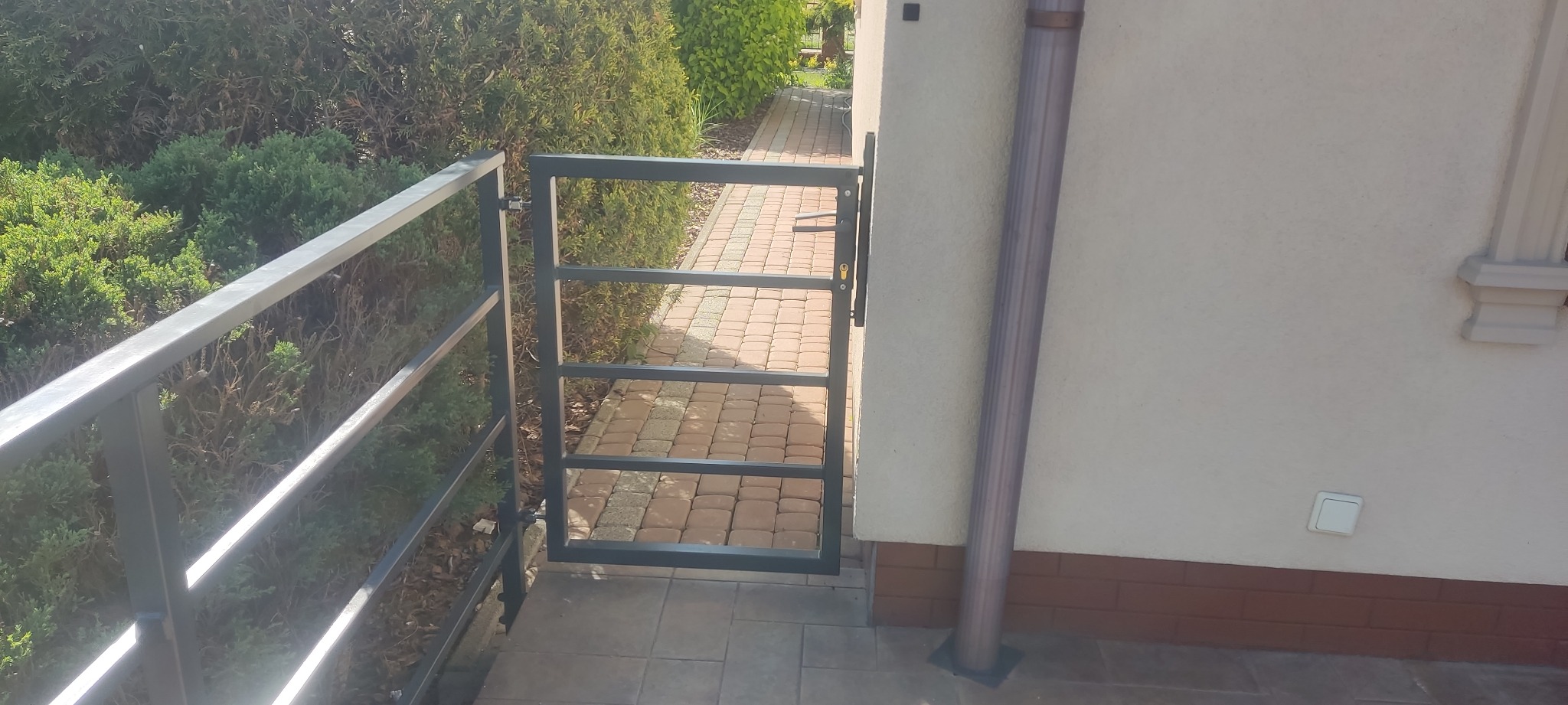 Metalowa furtka z poziomymi szczeblami, w kolorze grafitowym, zamontowana przy ogrodzeniu, z widocznym mechanizmem zamykania, na tle zieleni i ścieżki z kostki.