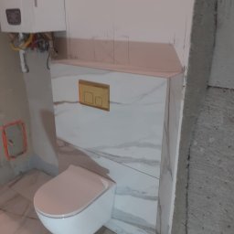 Kołobrzeska 6 - Podwieszana toaleta z białym marmurem, zabudowa stelaża z płyt karton-gips, widoczne ślady po ołówku na ścianie, w tle piec gazowy i surowy beton