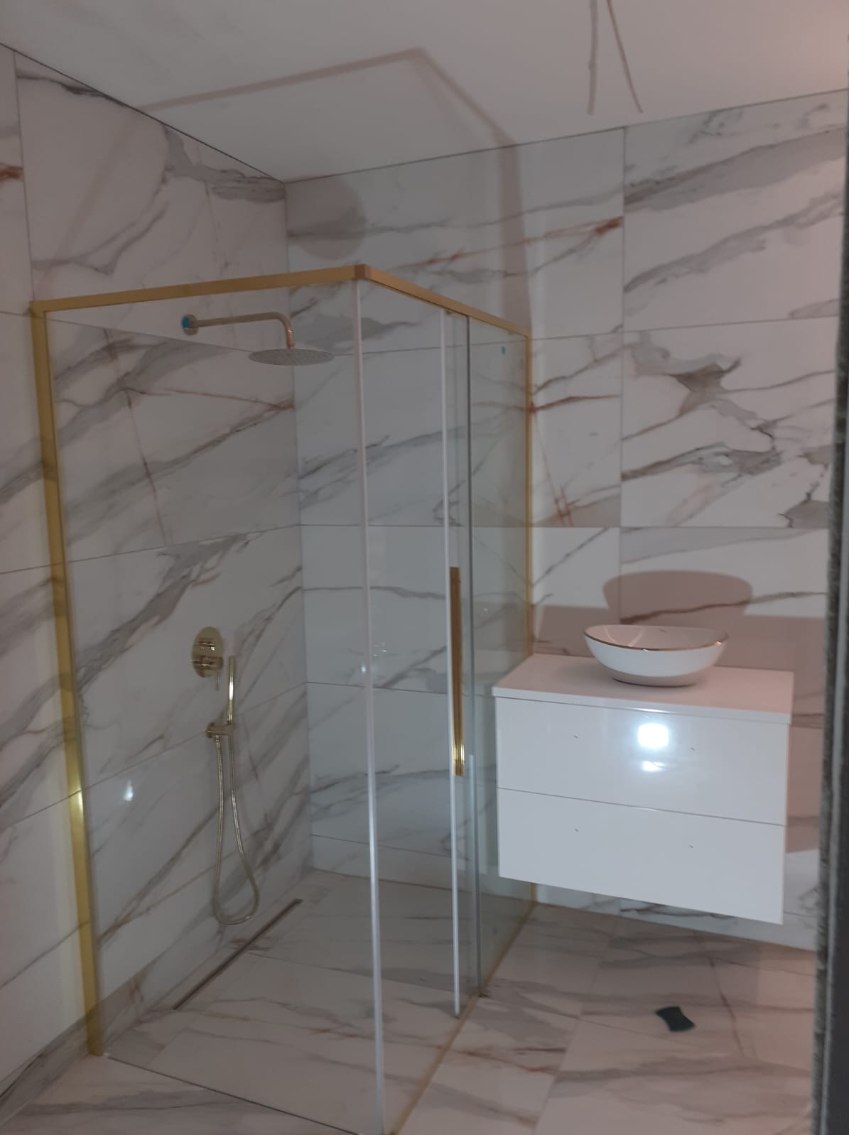 Nowoczesna kabina prysznicowa ze złotymi profilami na tle marmurowych płytek, biała szafka umywalkowa z misą w minimalistycznym stylu.