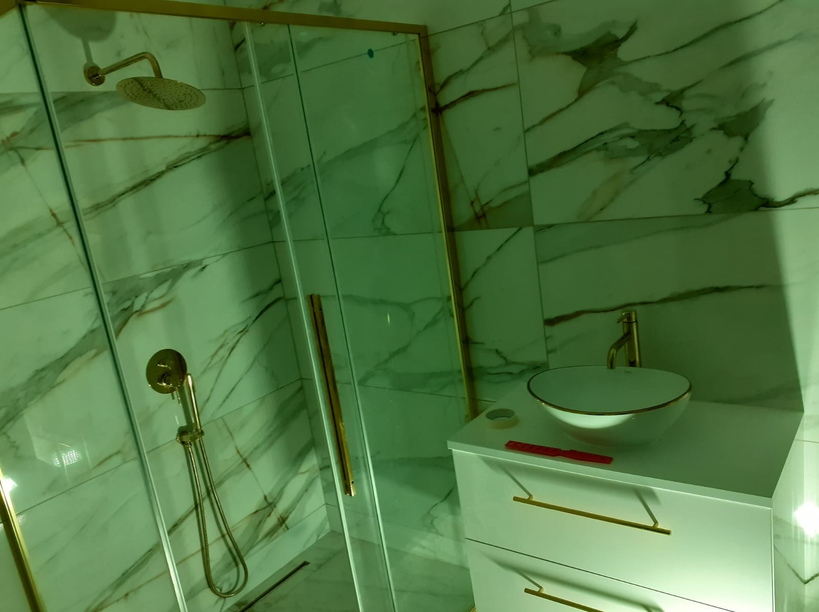 Nowoczesna łazienka z marmurowymi płytkami, złotymi akcentami na armaturze i kabinie prysznicowej, biała umywalka nablatowa z szafką, widoczna miarka i taśma malarska.