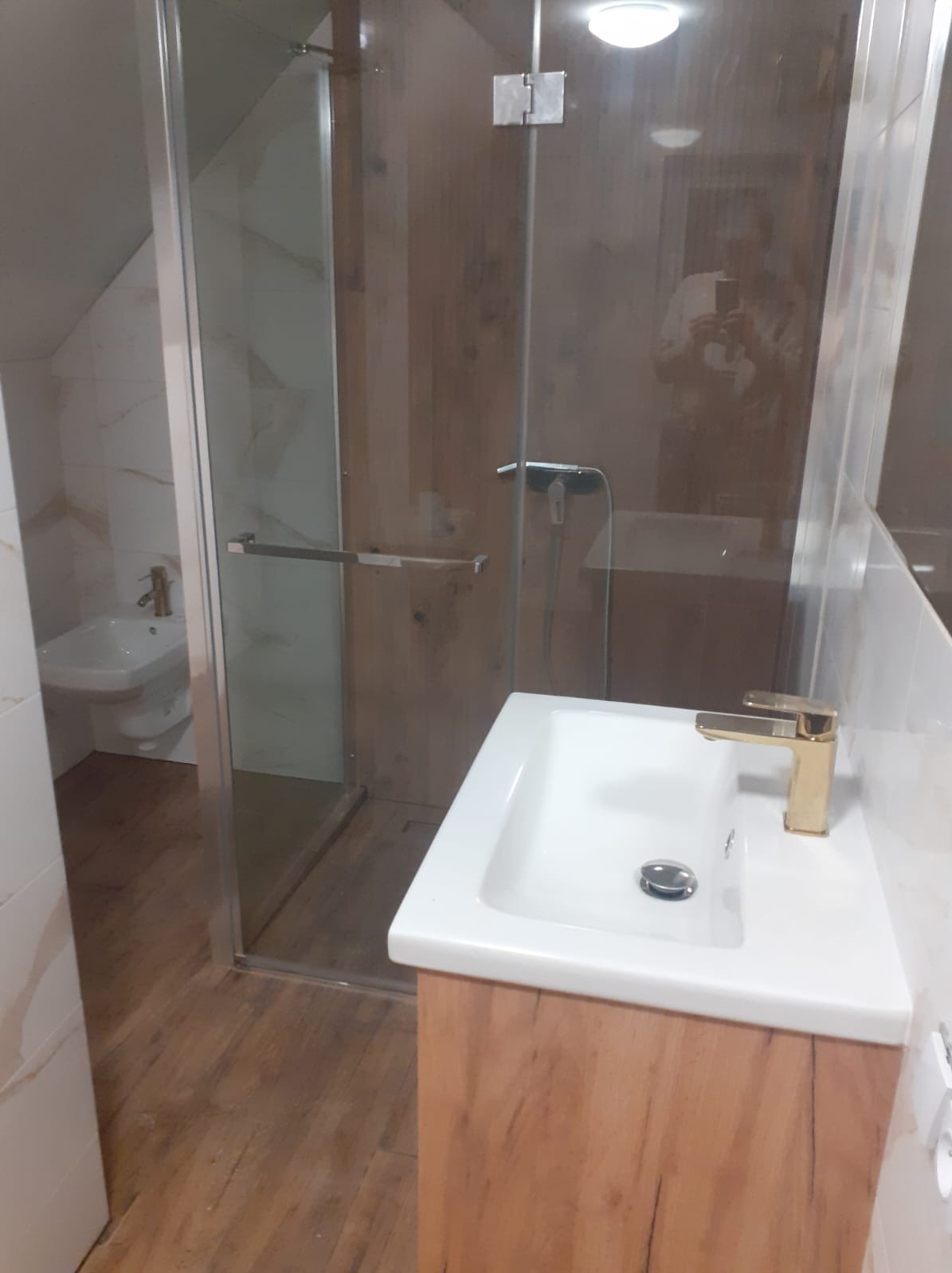 Umywalka z szafką o drewnianym wzorze i złotą baterią, kabina prysznicowa z ciemnego szkła z widocznym bidetem w tle, drewniana podłoga, białe kafelki częściowo na ścianach.