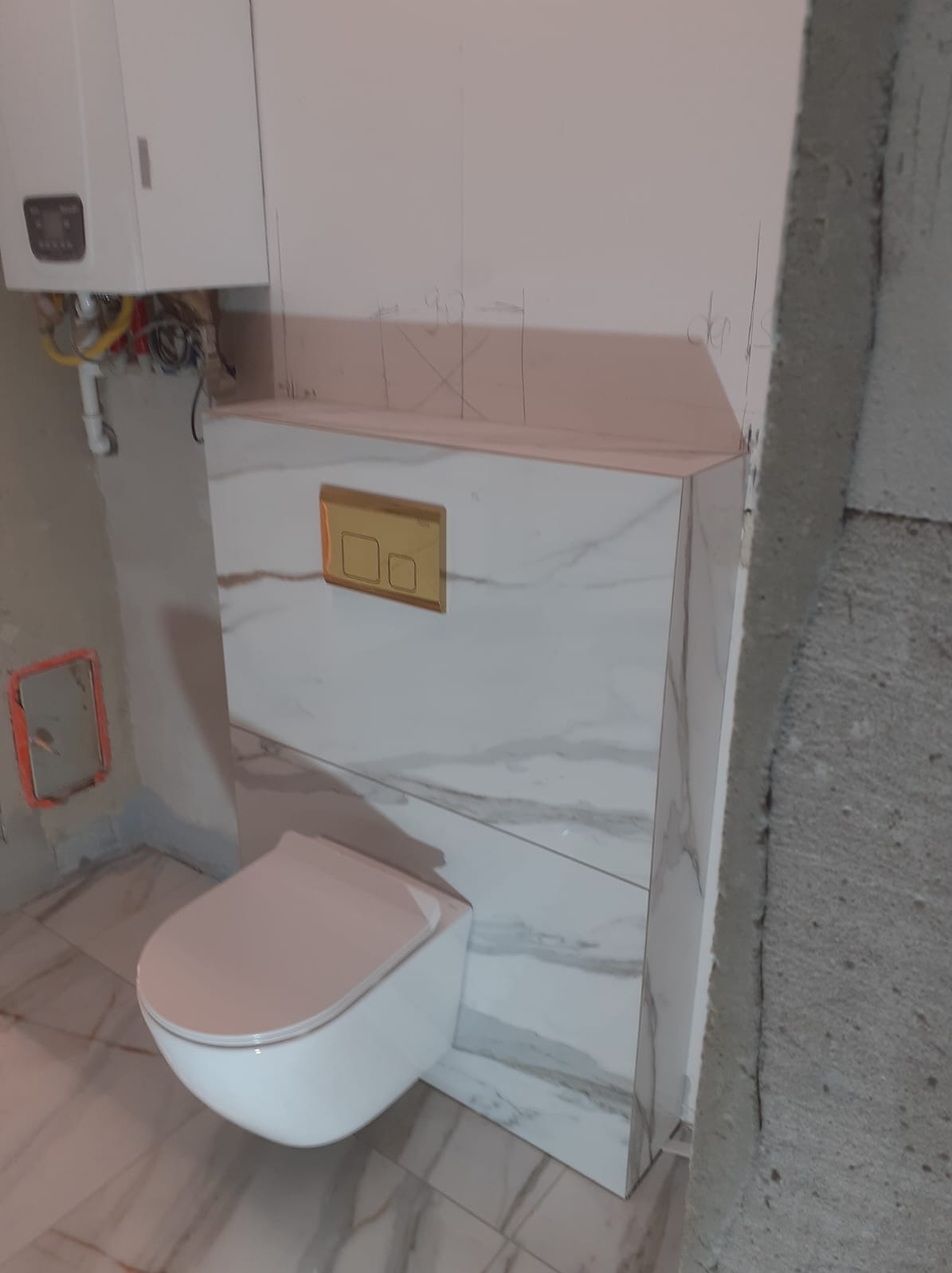Podwieszana toaleta z białym marmurem, zabudowa stelaża z płyt karton-gips, widoczne ślady po ołówku na ścianie, w tle piec gazowy i surowy beton