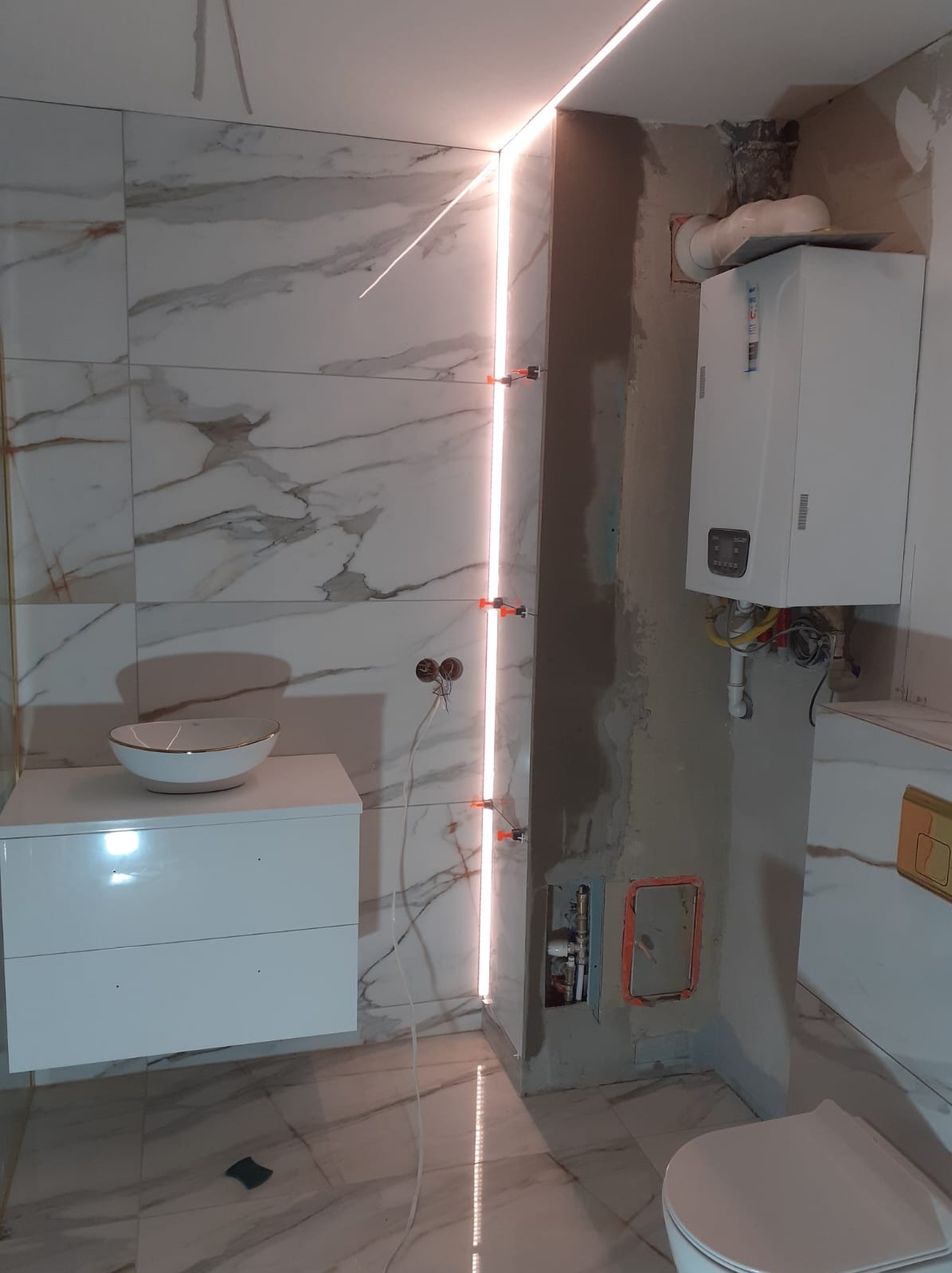 Łazienka w trakcie remontu: białe marmurowe płytki, podświetlenie LED, umywalka na szafce, piec gazowy, widoczne instalacje i poziomujące krzyżyki glazurnicze.